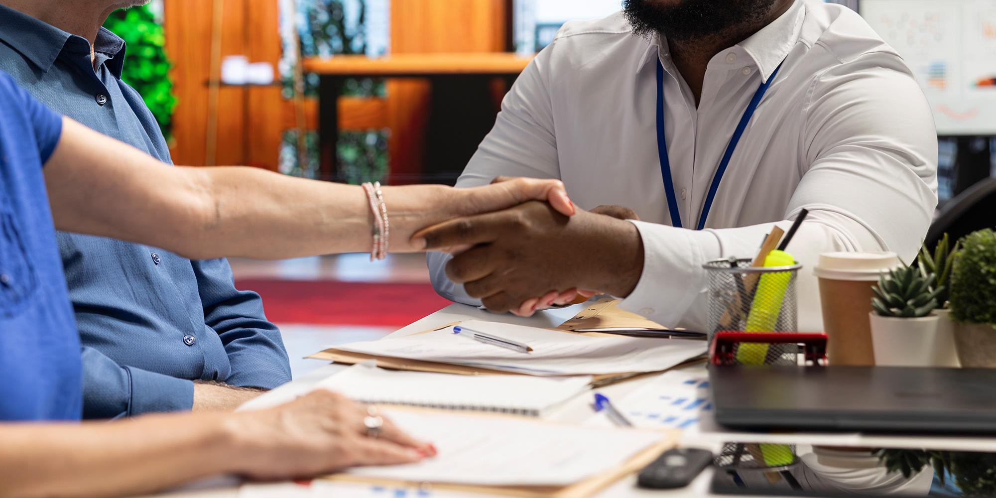 A Black man shakes a white woman's hand while another person sits nearby.